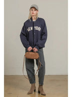 Woman wearing navy bomber jacket with New York lettering, grey cap, black jeans, and tan handbag and boots.