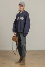 Woman wearing oversized navy bomber jacket with New York lettering, gray jeans, gray cap, and brown suede boots with bag