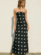 Woman wearing a black strapless maxi dress with large white polkadots, standing against a plain light wall.