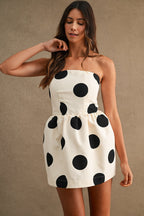 Woman wearing a strapless white mini dress with large black polkadots, styled with loose wavy hair against a plain background