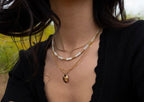 Close-up of a layered gold necklace with a pendant worn by a woman in a black top outdoors.