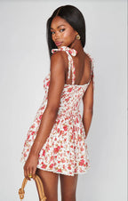 Woman wearing a floral romper with adjustable tie straps and smocked back, holding a wooden handbag in women's clothing style.