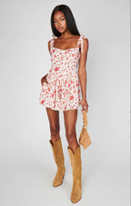 Woman wearing a floral romper with adjustable straps and pockets paired with tan suede cowboy boots and a matching bag