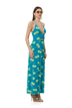 Woman wearing a teal maxi dress with green cherry print and gold accessories in a studio setting