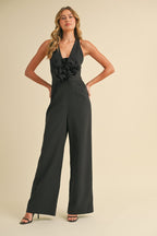 Woman wearing a black sleeveless wide-legged jumpsuit with a plunging neckline and large floral detail on the chest