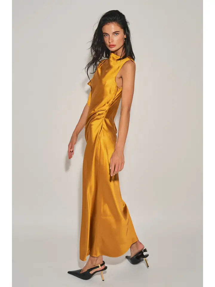 Woman wearing a golden satin maxi dress with an asymmetrical neckline and cap sleeve standing against a plain background.