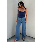 Woman wearing a navy scoop neck tank top paired with wide-leg blue jeans and black sandals standing against a plain wall.