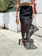 Woman wearing a black sheer overlay midi skirt with sandals, holding a small beige clutch outdoors on a sunny day