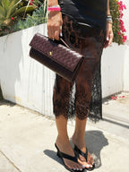 Black lace skirt with floral pattern worn with black wedge sandals and a dark woven clutch bag outdoors.