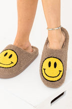 Brown fuzzy slippers with yellow smiley faces worn by a woman with a beaded ankle bracelet