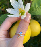 Woman’s hand wearing delicate gold snake ring with sparkling stones, holding white flower and yellow fruit outdoors