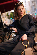 Woman wearing a dark pinstripe blouse sitting outdoors holding a beige handbag with gold hardware.