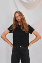 Woman wearing a midweight black tee paired with high-waisted black jeans, posing confidently against plain background.