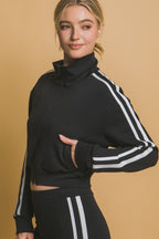 Woman wearing a black sporty jacket with white stripes on sleeves and matching high-waisted pants against tan background