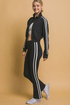 Smiling woman wearing black leggings with white side stripes and matching jacket, paired with white sneakers.