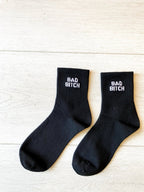 Pair of black women's socks with bold white text on the cuffs placed on a light wooden surface