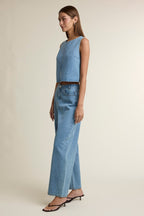 Woman wearing light blue denim pants and sleeveless cropped denim top with black heeled sandals against neutral background