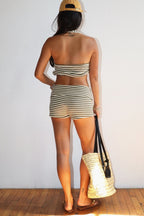 Woman wearing striped halter top and matching shorts, holding woven tote bag, standing on wooden floor.