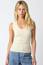 Woman wearing a beige ribbed sleeveless top with a U scoop neckline paired with blue jeans standing against a white background