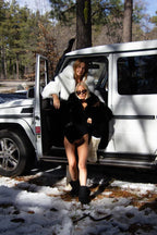 Two women wearing cropped faux fur jackets pose by a white SUV in a snowy forest setting