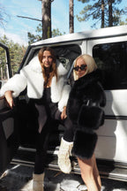 Two women wearing cropped faux fur jackets in white and black standing beside a white SUV outdoors in winter.