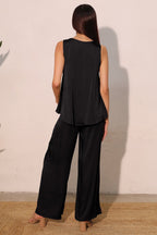 Woman wearing loose black sleeveless top and wide-leg pants standing against a pale wall in casual women's clothing.