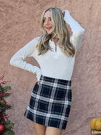 Woman wearing a black and white plaid mini skirt with a long-sleeve white ribbed top standing against a textured wall