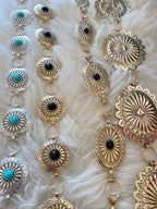Close-up of gold and silver metal belts with turquoise and black stone accents on a soft fabric background
