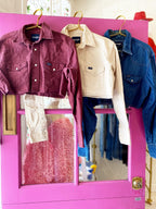 Three cropped western-style button-down shirts in burgundy, cream, and denim blue hanging on a pink door.