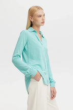 Woman wearing a sea-colored lightweight blouse with long sleeves and a collar, styled with white pants.
