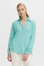 Woman wearing a sea-colored button-down blouse with whipstitch details, styled with white pants.