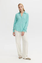 Woman wearing a sea-colored casual button-down blouse with whipstitch details and white wide-leg pants.