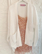 White oversized knit cardigan with pockets layered over a floral patterned dress on a hanger against a textured wall