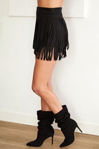 Woman wearing a black faux suede fringe skirt paired with black slouchy heeled boots against a white wall