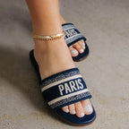 Woman wearing navy blue slip-on flat sandals with fabric straps embroidered with "PARIS" text