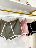 Three pairs of women's ribbed boy shorts in olive, pink, and black hanging on gold and white hangers.