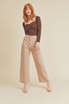 Woman wearing beige wide leg pants and a fitted dark brown long-sleeve top standing against a plain background